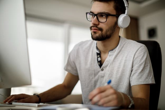 Homem sentado em frente ao computador com headphones enquanto anota algo em caderno