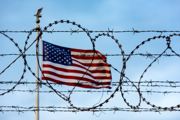 saiba o que fazer e como sair dos eua com o visto americano vencido foto de arame farpado com bandeira dos eua no fundo sinalizando dificuldades com visto americano vencido